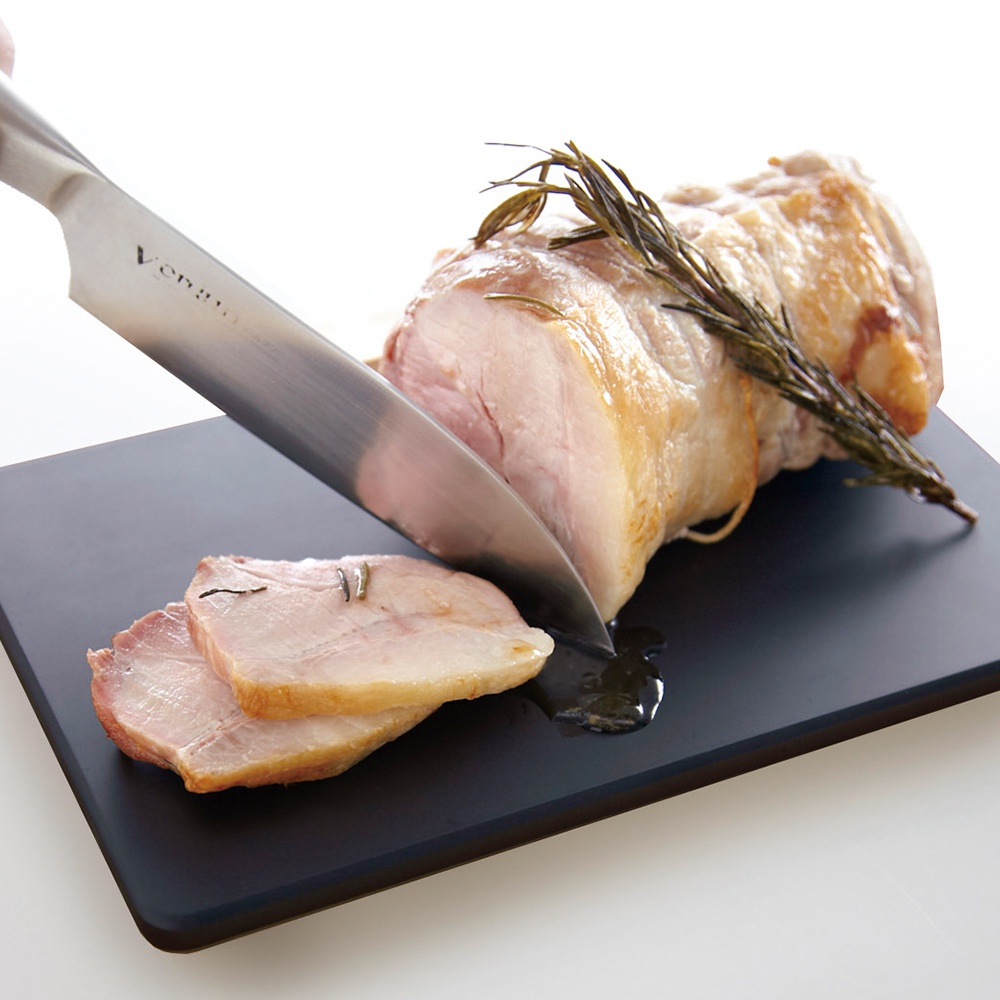 一個黑色砧板上，一把銀色金屬刀正在切一片烤肉，烤肉上裝飾著迷迭香，烤肉旁有兩片切好的烤肉。