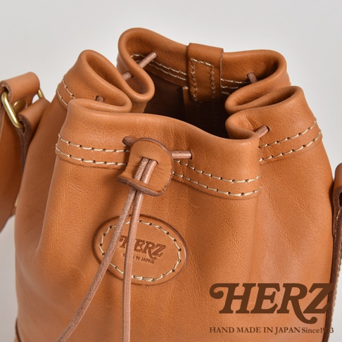 一個棕色皮革束口單肩包，有細緻的縫線細節，包身中央有一個橢圓形壓印的品牌標誌，束口處有皮革拉繩。