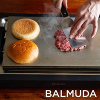 黑色極致電烤盤上，兩顆漢堡麵包旁，有人用金屬鍋鏟將生絞肉壓平在烤盤上，烤盤冒出蒸氣。