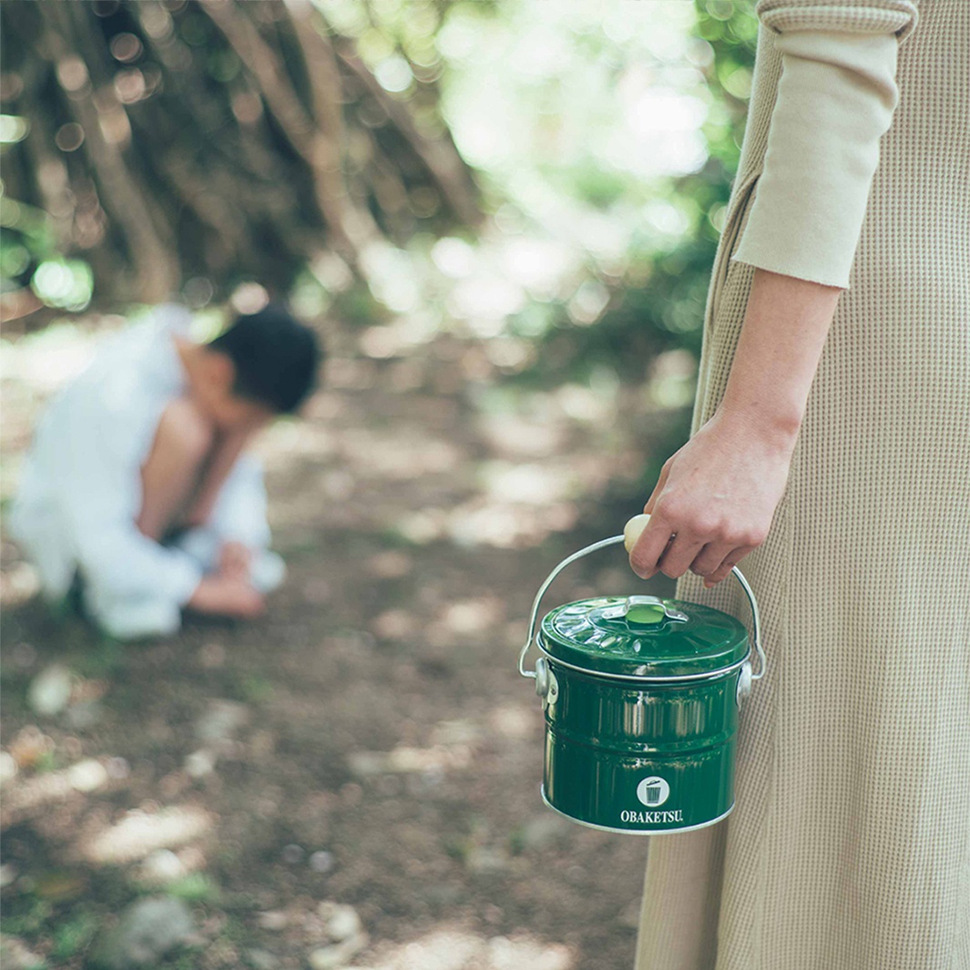 綠色OBAKETSU蚊香桶，有提把和蓋子，上面有白色標誌，背景是模糊的樹林和一個坐在地上的孩子