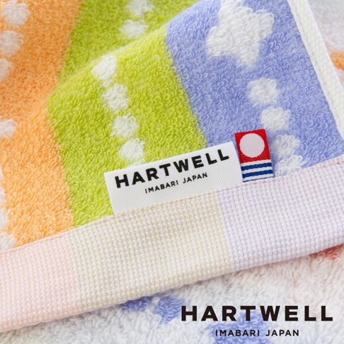 這款 Hartwell 今治森林動物刺繡浴巾採用柔軟的毛圈織物製成，表面呈現橙、綠與紫色的繽紛條紋圖樣，並綴有白色圓點細節。浴巾邊緣縫有一枚印有 Hartwell Imabari Japan 字樣與紅白色圓形圖標的精緻布標，整體質感細膩柔和。
