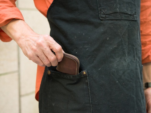 棕色皮革製的拉鍊式零錢包，由手放入黑色圍裙口袋中。