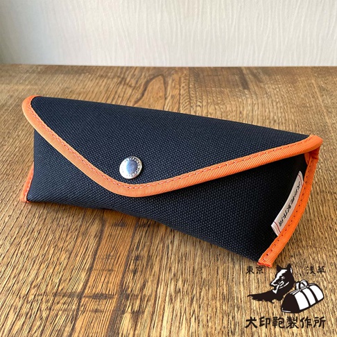 一個藍色帆布製的眼鏡盒，橙色滾邊，銀色按扣，上面有犬印鞄製作所的標誌。