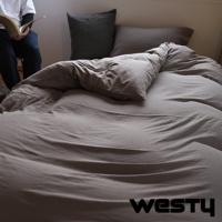 寬敞的床鋪鋪著灰色床單，上面堆疊著柔軟的被子和枕頭，床邊坐著一個人正在閱讀。