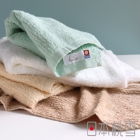 疊放的今治細絨浴巾，包含淺綠色、白色、米色和褐色。淺綠色浴巾的邊緣有一個標籤，上面印有「hiorie」字樣和「MADE IN JAPAN」的字樣。