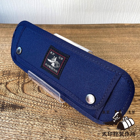 一個深藍色純棉帆布製成的長方形收納包，表面縫有黑色標籤，標籤上有日文圖案和文字。收納包側邊有金屬釦，整體呈現堅固耐用的質感。