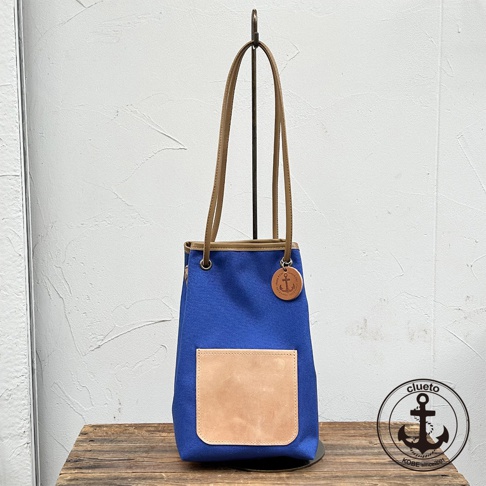 神戶clueto品牌藍色防潑水尼龍束口肩背包，配有義大利皮革口袋和船錨吊飾，置於木製桌面上。