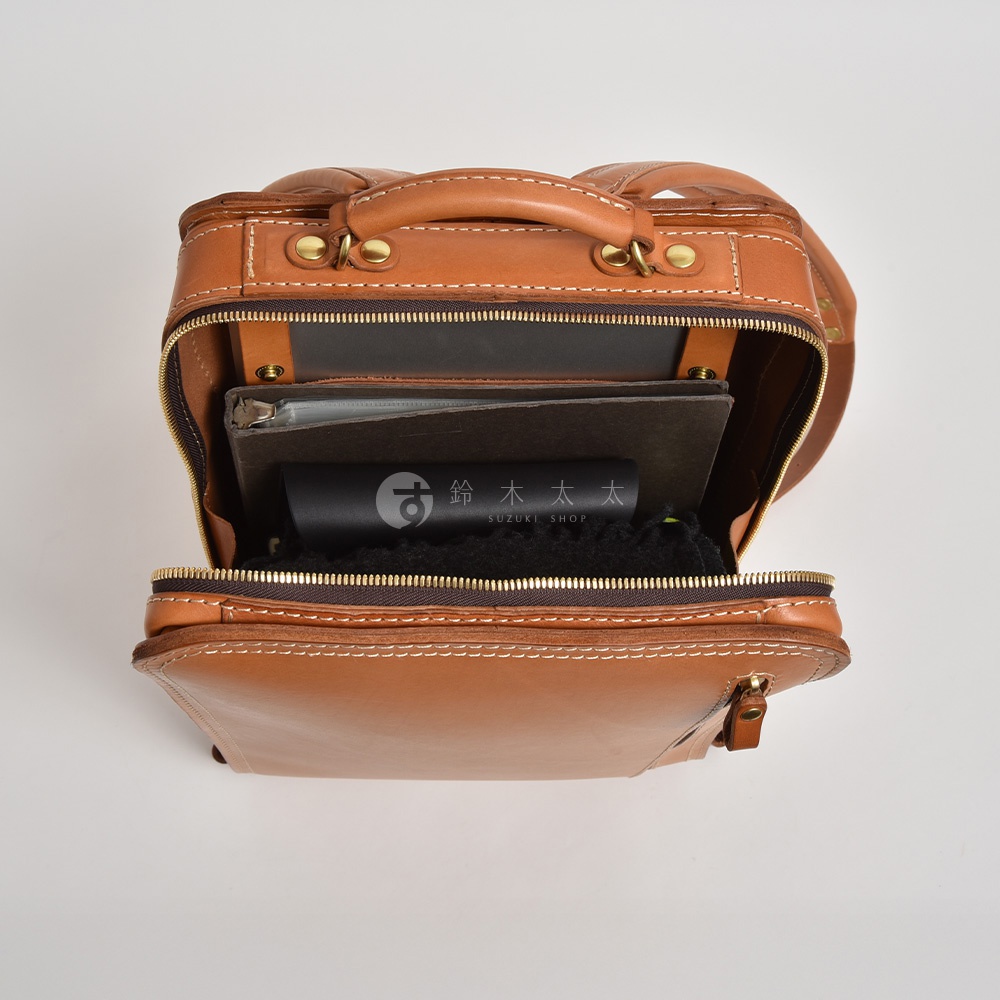 職人手作皮革商務電腦後背包，呈現自然棕色調，以精緻縫線裝飾。包款頂部有提把，主隔層打開，內有文件夾、筆記本及其他物品，材質顯現皮革質感。