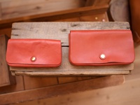 兩款橘紅色皮革長夾，皆配有金色按扣。左邊較小，右邊較大，均置於木製層架上。