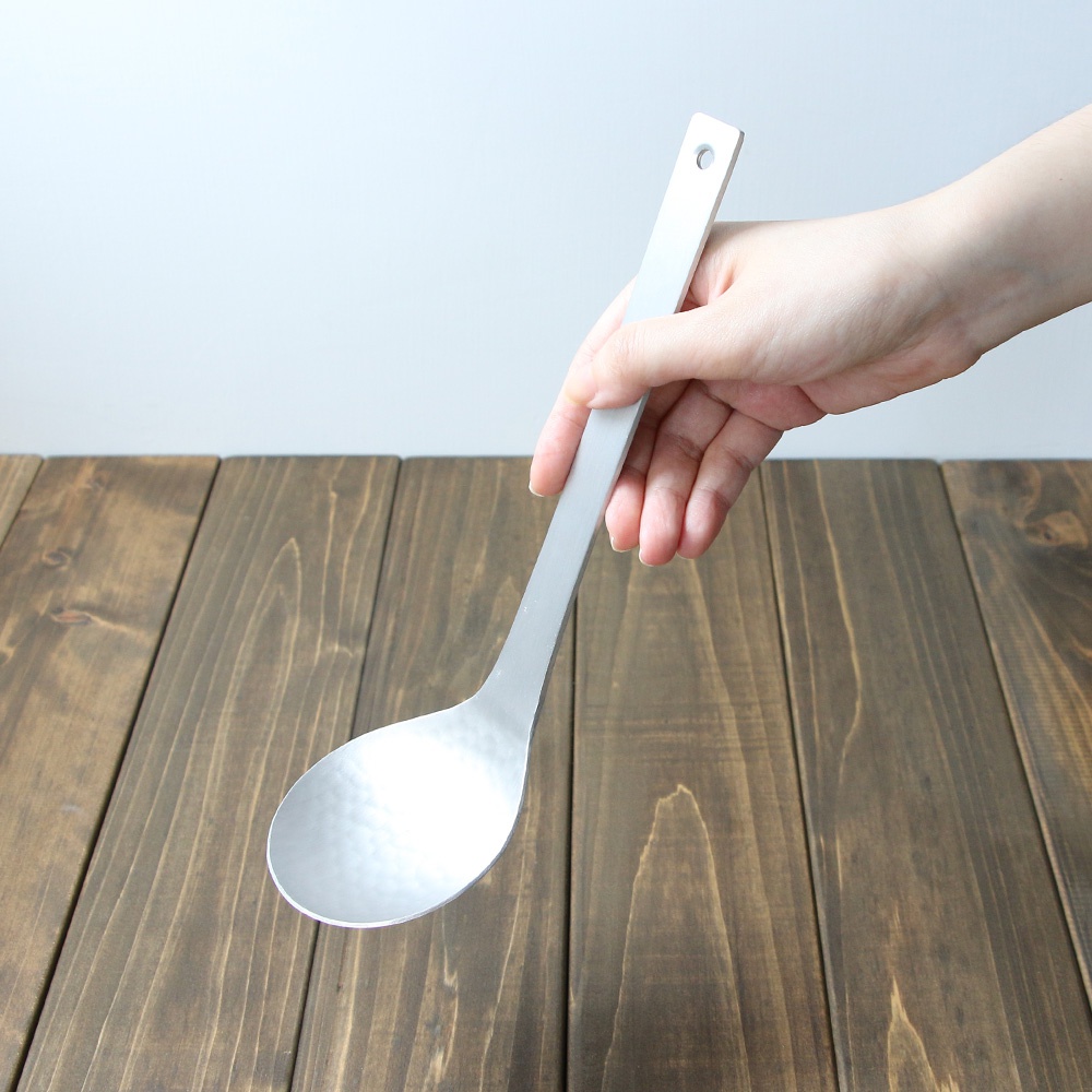 一只手正拿著一個銀色鋁製湯勺，湯勺的勺面帶有錘目紋。