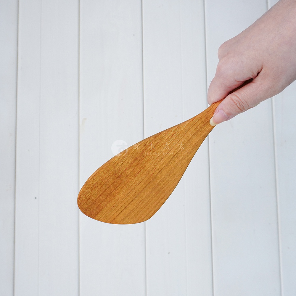 一隻手正拿著一把淺棕色、木質紋理清晰的山櫻木斜口鍋鏟，鍋鏟邊緣略帶弧度，適合煎炒使用。