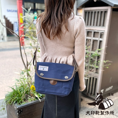 一名女性肩背著一個深藍色純棉帆布郵差包，包蓋上有棕色皮革飾邊和兩個銀色金屬扣。