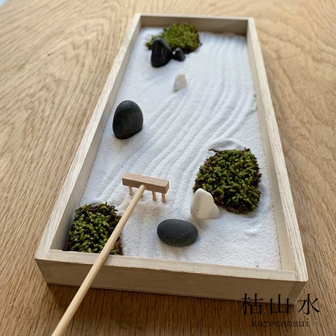 日式枯山水模型，淺木色方形底座，鋪滿白色細沙，沙上耙出波浪紋路。點綴著三塊深灰色鵝卵石、兩塊白色鵝卵石，以及三叢翠綠色苔蘚。一迷你木製耙子放置於沙盤一側。