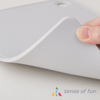 淺灰色矽膠砧板，材質柔軟，邊緣圓潤。一隻手正在彎曲砧板的一角，展示其彈性。砧板表面帶有細微紋理，右上角有一個圓形掛孔。