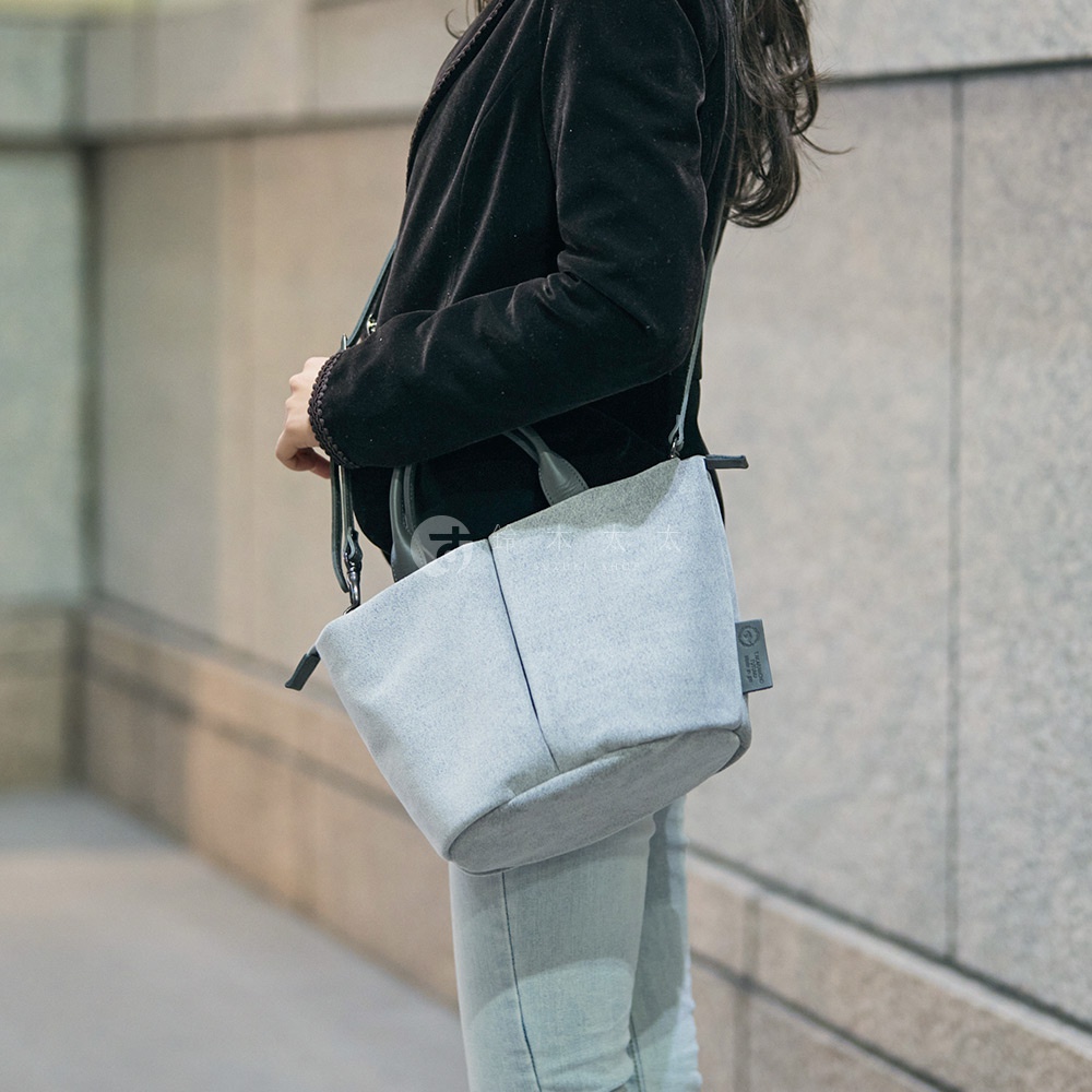 一名身穿黑色外套和淺色牛仔褲的女子，斜挎著一個淺灰色的TUTUMU Suede Marche petit 兩用單肩包。