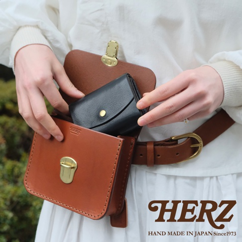 職人手作皮革經典腰包，使用全皮革製作，主體為焦糖棕色，內側放入一個黑色短夾，腰包以金色鎖扣開合，搭配棕色皮革腰帶，品牌標誌印於腰帶扣旁。