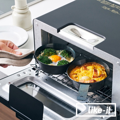 兩個黑色鐵製圓形調理鍋，裝有烤雞蛋和花椰菜，以及培根烘蛋，置於烤箱架上。其中一個鍋子由戴著隔熱手套的手握住。