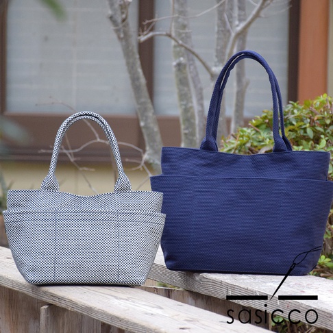 兩款三河木綿刺子織手提托特包，一款為白色底黑格紋，另一款為深藍色。兩款皆為 L 尺寸，採用經典托特包款式，有雙提把，包身呈現樸實的織紋質感，外側設有方便收納的口袋。