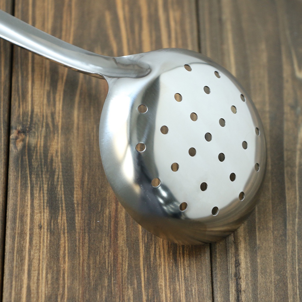 一個不鏽鋼中型撈勺，帶有圓形孔洞，放在木紋桌面上。
