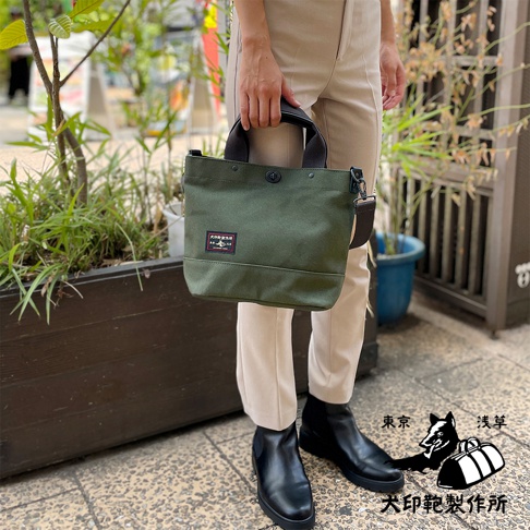 綠色帆布材質的兩用托特包，有黑色提把與可拆卸斜背帶，包身正面有兩個方形布貼，其中一個印有黑犬圖案。