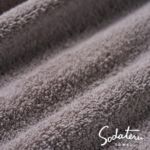 特寫畫面，展示一條淺灰色、蓬鬆柔軟的毛巾，其織物紋理清晰可見，並帶有品牌標誌 Sodateru TOWEL。