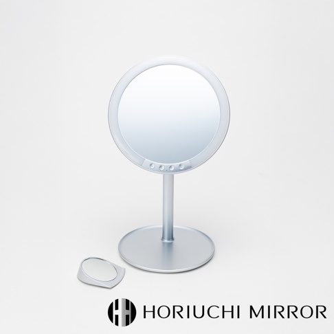 銀色金屬材質的桌上型化妝鏡，鏡面為圓形，鏡框下方有數個圓形觸控按鈕。鏡子底部為圓形底座，帶有支撐桿。一旁有一個小型的三角形放大鏡。