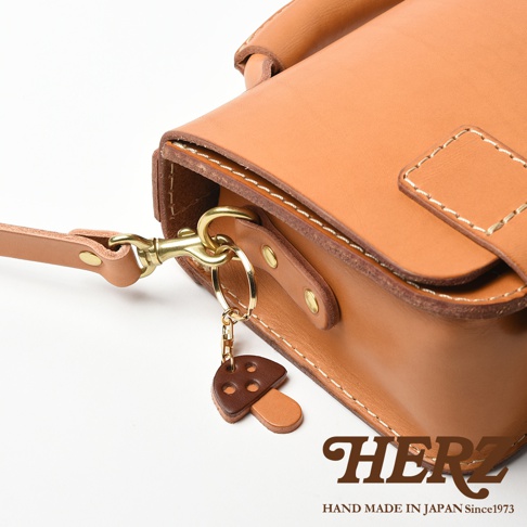 職人手作皮革香菇造型鑰匙圈，由兩塊淺棕色皮革組成香菇造型，傘蓋上方有三個深棕色皮革圓點裝飾，搭配金色金屬掛環與鉤扣，掛於淺棕色皮革包的提把上。