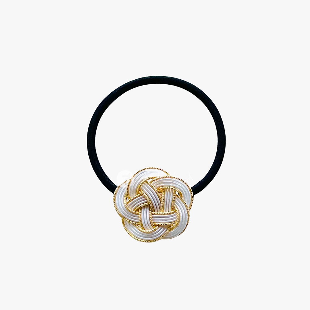 一個黑色彈性髮圈，配有一個精緻的日式水引花結裝飾，由白色和金色的細線交織而成，形成複雜的編織圖案。