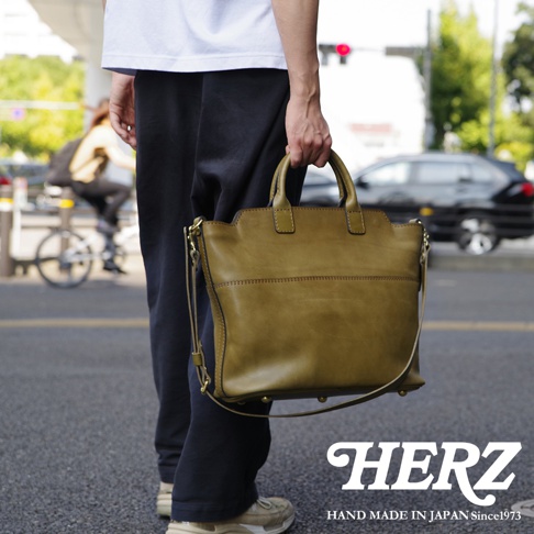 職人手作皮革兩用托特包，橄欖綠，方形結構，頂部有提把，附可拆卸肩帶，包身有縫線細節與金屬腳釘。