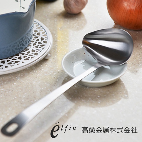 日製不鏽鋼橢圓尖嘴湯杓，湯勺呈銀色金屬光澤，置於白色陶瓷醬料碟中，湯勺柄上有掛孔，背景為淺色桌面，有洋蔥和蒜頭。