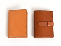兩款職人手作皮革卡片收納夾，左邊為淺棕色，右邊為深棕色，皆為對開式設計，並以白色縫線車邊，右側收納夾配有皮革束帶。