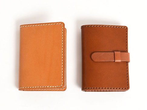 兩款職人手作皮革卡片收納夾，左邊為淺棕色，右邊為深棕色，皆為對開式設計，並以白色縫線車邊，右側收納夾配有皮革束帶。