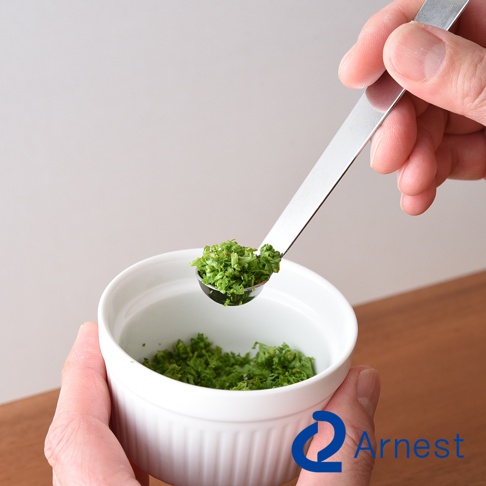 一只手拿著不鏽鋼萬用醬料小勺，勺子裡裝有切碎的綠色香草，正準備放入一個白色陶瓷碗中。碗裡也裝有切碎的香草。