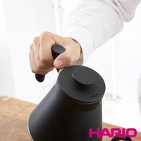 一只手正握著霧黑色細口手沖壺的把手，壺身為啞光黑色，壺蓋上有三個小圓孔。