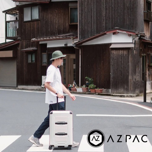 一位男子在日式建築前拉著一個米白色 26 吋旅行箱，箱子上有銀色拉鍊和黑色條紋裝飾。