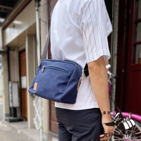 一位身穿條紋短袖上衣的男子，側背一個深藍色帆布斜背包，包包上有一個淺棕色的皮革標籤。