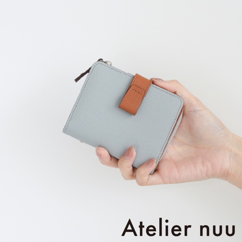 一隻手拿著一個灰色荔枝紋皮革製成的 Atelier nuu 雙折短夾，短夾中央配有棕色皮革扣帶裝飾，上方帶有精緻的銀色拉鍊細節，呈現簡約優雅的質感。