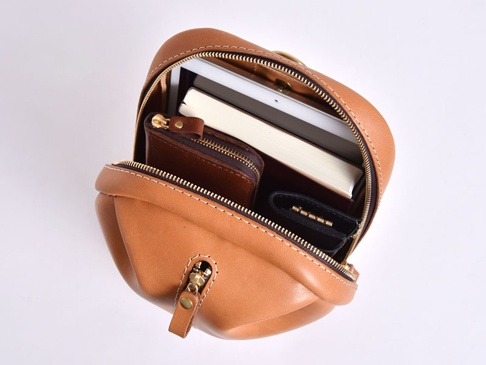 職人手作皮革立體運動斜背包，以棕褐色皮革製成，搭配金色拉鍊。包內可見一本白色封面的書、一個咖啡色皮革零錢包，以及一個黑色鑰匙包。