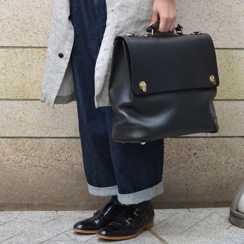 黑色皮革波士頓包，配有金色搭扣和手柄，由一位身穿灰色外套和牛仔褲的人士攜帶。