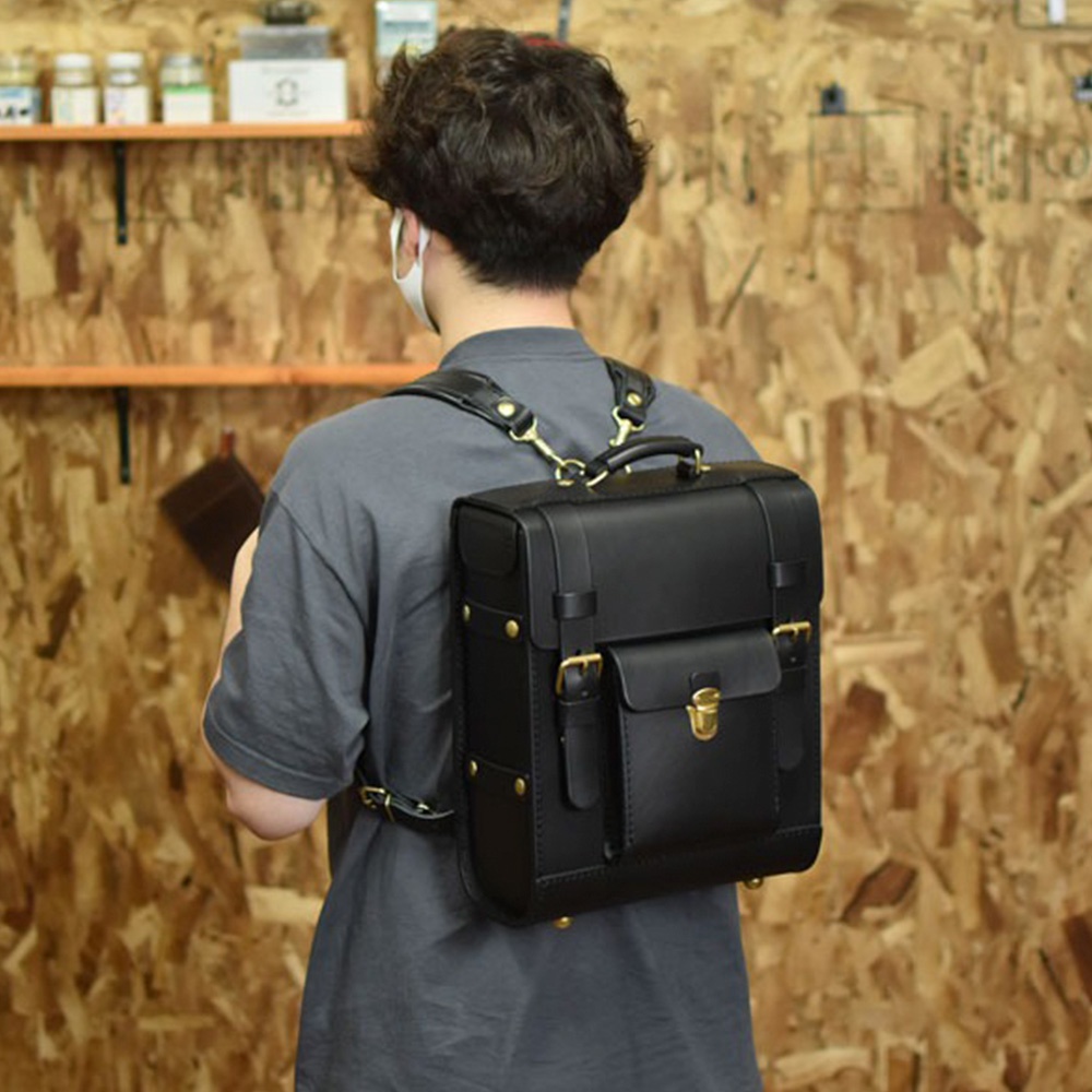 黑色皮革學院風後背包，配有前置翻蓋口袋、金色金屬鎖扣、可調節肩帶及頂部提把。