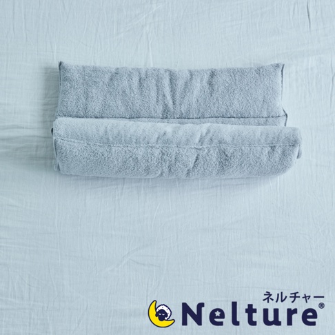 淺灰色毛巾材質的捲捲枕，柔軟蓬鬆，呈現出雙層捲繞的設計，放在淺藍色床單上。