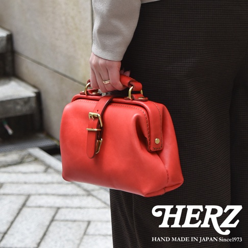 一位女性手持紅色皮革迷你軟皮杜勒斯包，包款採用經典醫生包造型，有皮質提把、金屬釦環與皮革束帶，整體風格復古簡約。