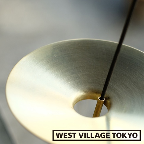 黃銅製的線香架，圓形盤狀設計，中央有一個圓孔，一根細長的黑色線香從孔中穿過，線香架表面呈現拉絲質感，光澤溫潤。