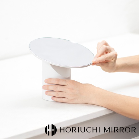 白色桌上型可調節化妝圓鏡，鏡面為銀色，底座為圓柱形白色，由雙手調整鏡面角度。