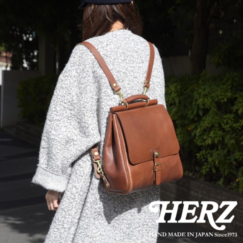 一位身穿灰色毛衣的女性背著一個棕色皮革後背包，包包採用屋瓦設計，配有可調節的肩帶和金屬扣環。