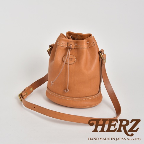 棕色皮革束口單肩包，圓筒狀包身，頂部束繩設計，配有可調節長度的皮革背帶，包身中央有圓形皮革品牌標誌。