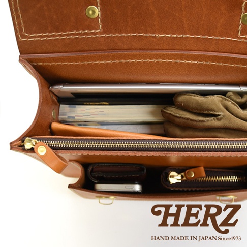 職人手作皮革經典雙扣兩用單肩包，內部展示，包含平板電腦、書籍、手套、錢包和手機。包包為棕色皮革材質，配有黃銅色拉鍊和鈕扣。