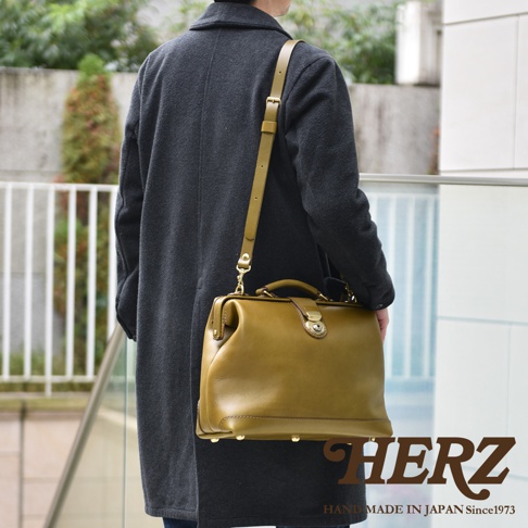 一位穿著深灰色長大衣的男性，肩背著一個橄欖綠色的日本HERZ手工皮革杜勒斯公事包，包款採用經典醫生包設計並配有金屬扣環與底部金屬腳釘，整體呈現沉穩簡約的質感。