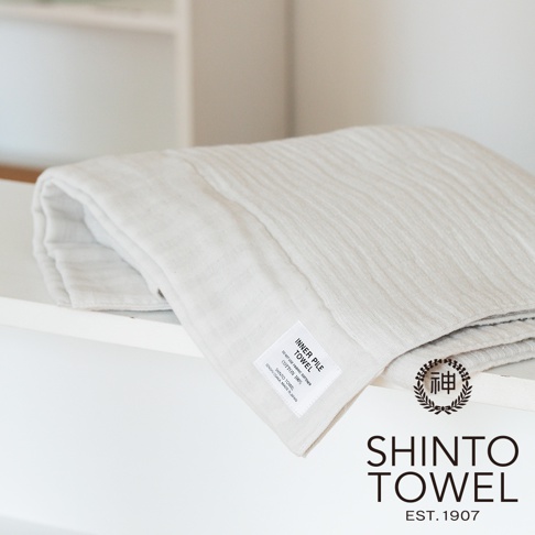 一條淺米色雙面紗布輕質毛巾，材質為純棉，表面有細緻的紋理，附有產品標籤。