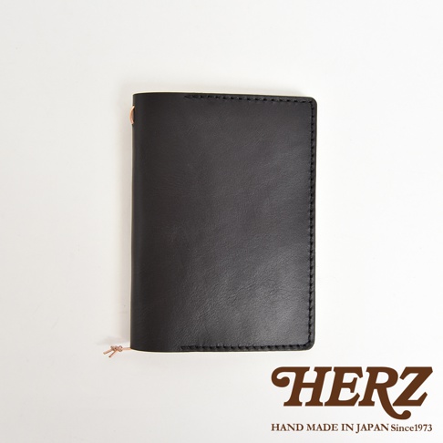 黑色皮革手帳書套，有精緻縫線細節，底部有銅色的小掛飾，下方印有HERZ品牌字樣和HAND MADE IN JAPAN Since1973字樣。